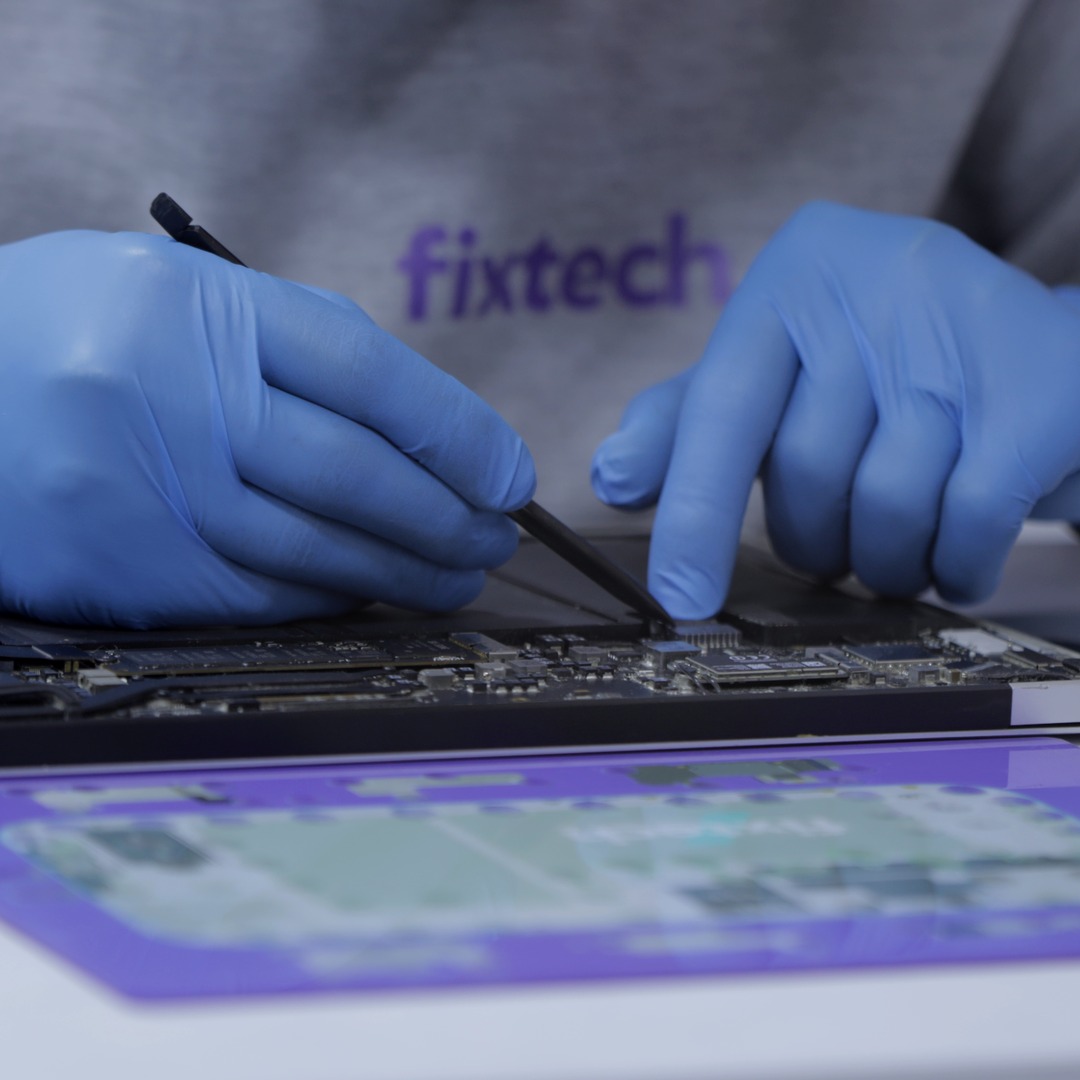 Gloved hands working on an open MacBook logic board with a fine tool, over a Fixtech mat.